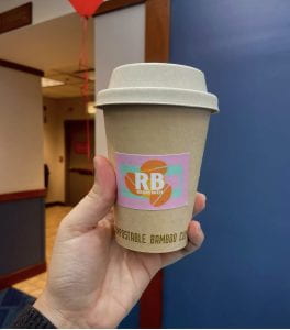 A ReBrewed coffee cup. It is medium-sized with a bright, modern logo made up of the letters RB super imposed on brightly colored coffee beans