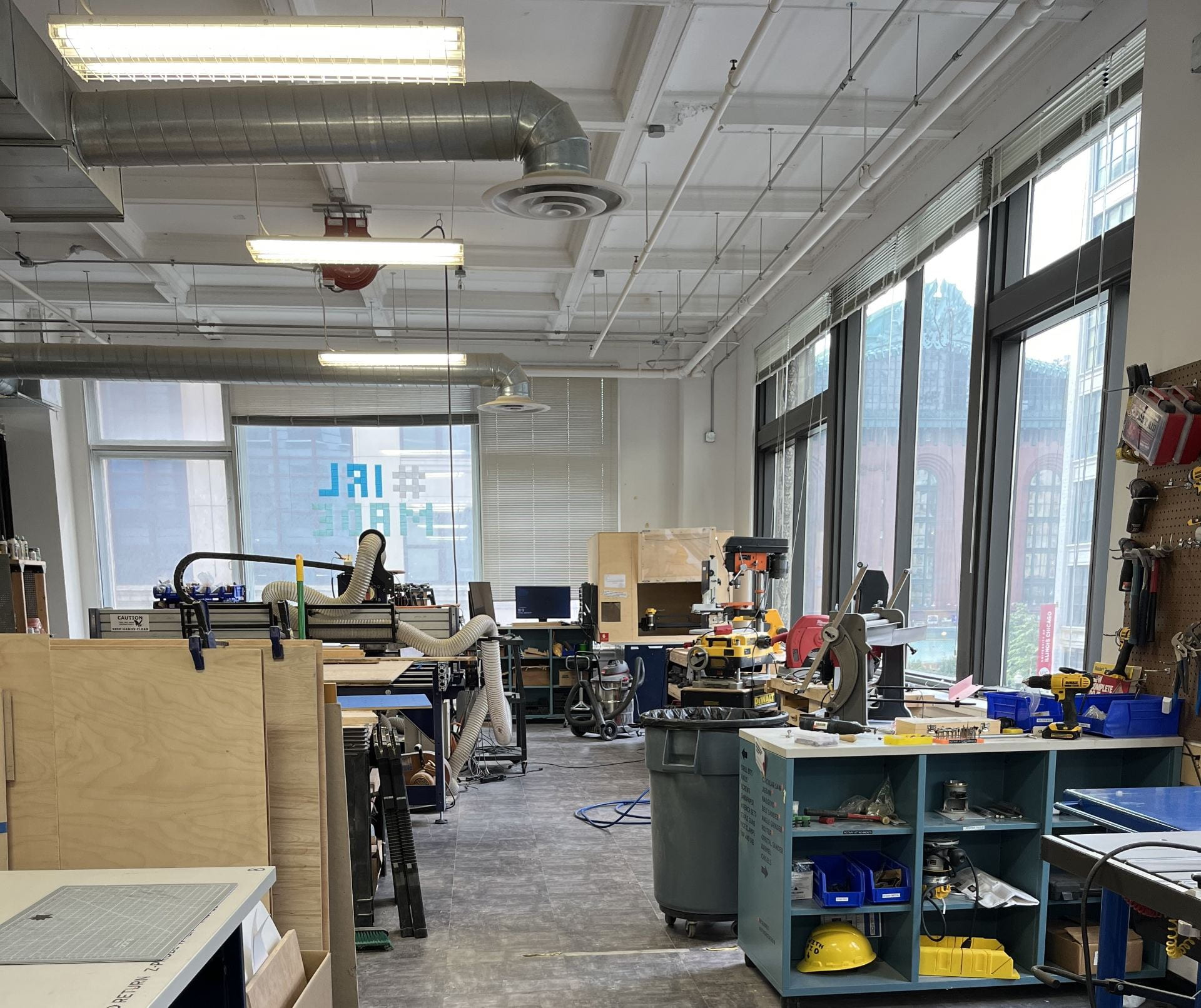 A big room with lots of woodworking tools, benches, and pieces of wood, with windows showing neighboring Chicago buildings.