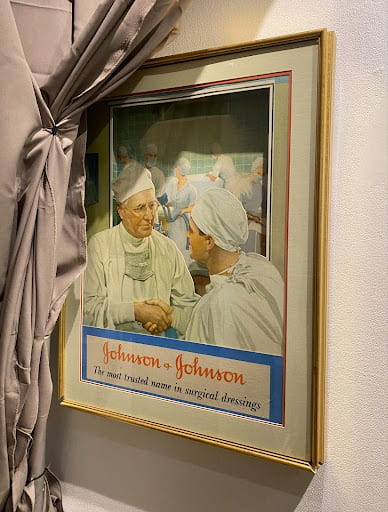 A framed illustration of two surgeons, just on the other side of the operating, shaking hands. Text at the bottom reads "Johnson & Johnson: the most trusted name in surgical dressings."