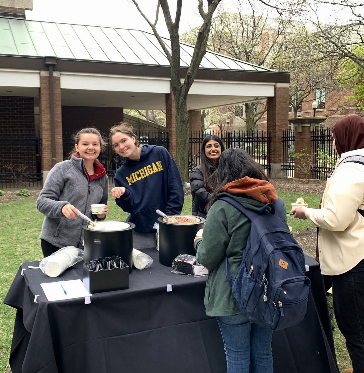 A group of Pathways Honors students serving themselves ice cream at an outdoor table in a courtyard. Two students face the camera and smile.