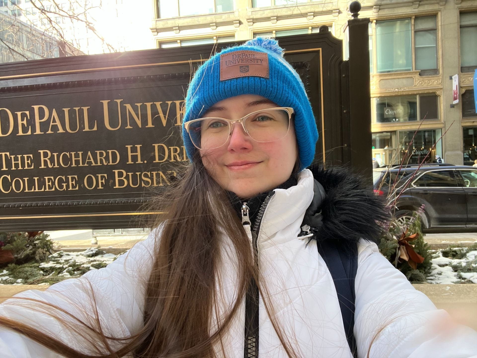 Sienna standing in front of the DePaul University, Driehaus College of Business sign, with a DePaul Blue Beanie on.