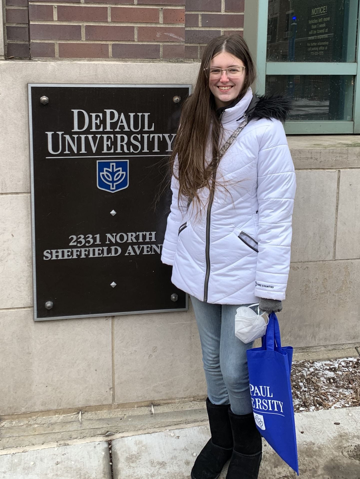 Sienna smiling in front of a DePaul University Sign, in a white long winter coat in winter.