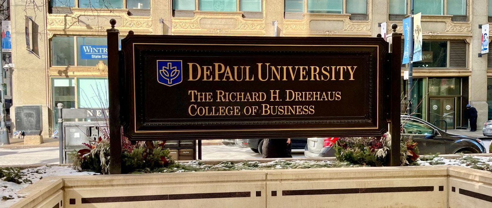 A black sign outside with snow on the ground with the words “DePaul University” “The Richard H. Driehaus College of Business” in blue and gold colors.