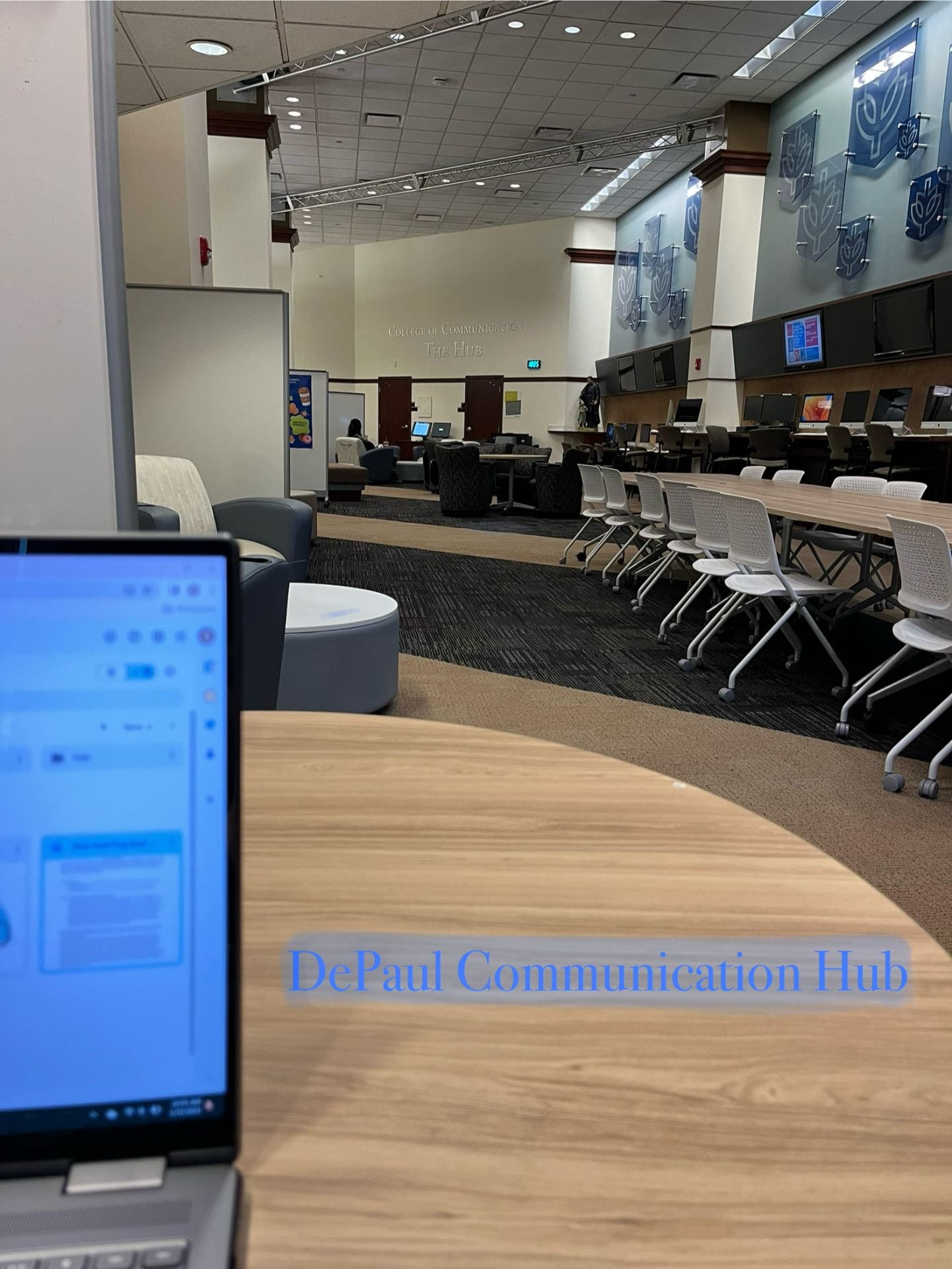 A laptop open on the edge of the screen, facing out lots of tables, computers, and comfy chairs in a computer lounge located at DePaul. With the words “DePaul’s Communication Hub” in blue at the bottom. 