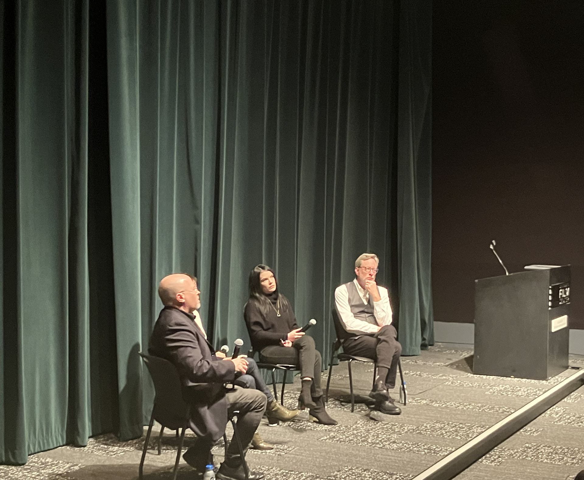 A panel of sitting people in front of a theatre curtain. They are taking questions from the audience.