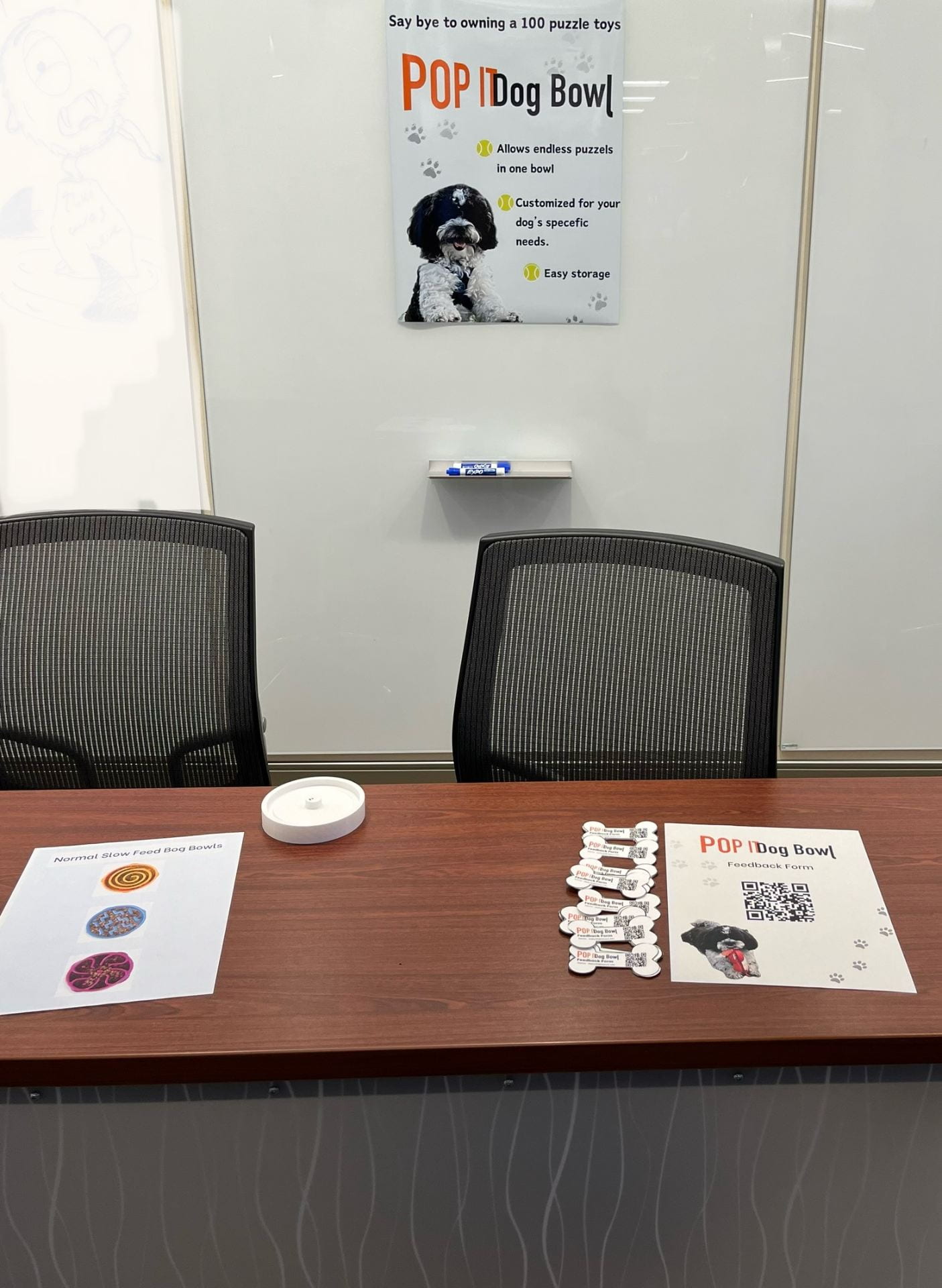 A poster hanging with the words “Popit Dog Bowl” in bold with a picture of a dog on it. The table has a flier and little paper dog bones with a QR code on it.