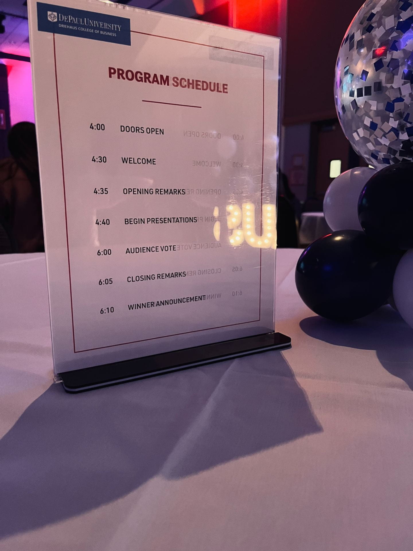A white table with a depaul blue balloon centerpiece. Along with a program schedule outline the Driehaus Cup schedule.