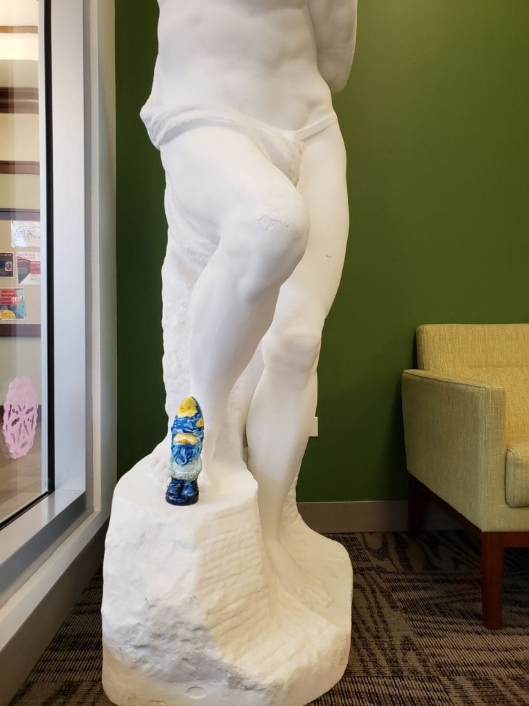 a small garden gnome painted to resemble Vincent van Gogh's Starry Night standing on a copy of a recreation of Defiant Prisoner by Michelangelo