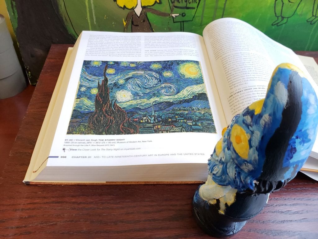 a small garden gnome painted to resemble Vincent van Gogh's Starry Night standing as if looking at a picture of Starry Night in a book