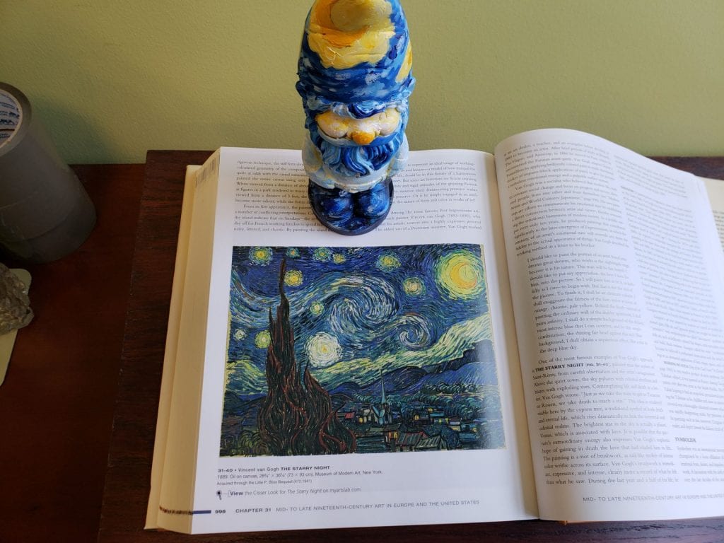 a small garden gnome painted to resemble Vincent van Gogh's Starry Night standing near a picture of Starry Night in a book 