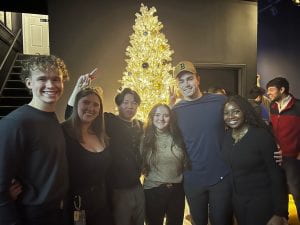 A group of five students poses playfully in front of a christmas tree