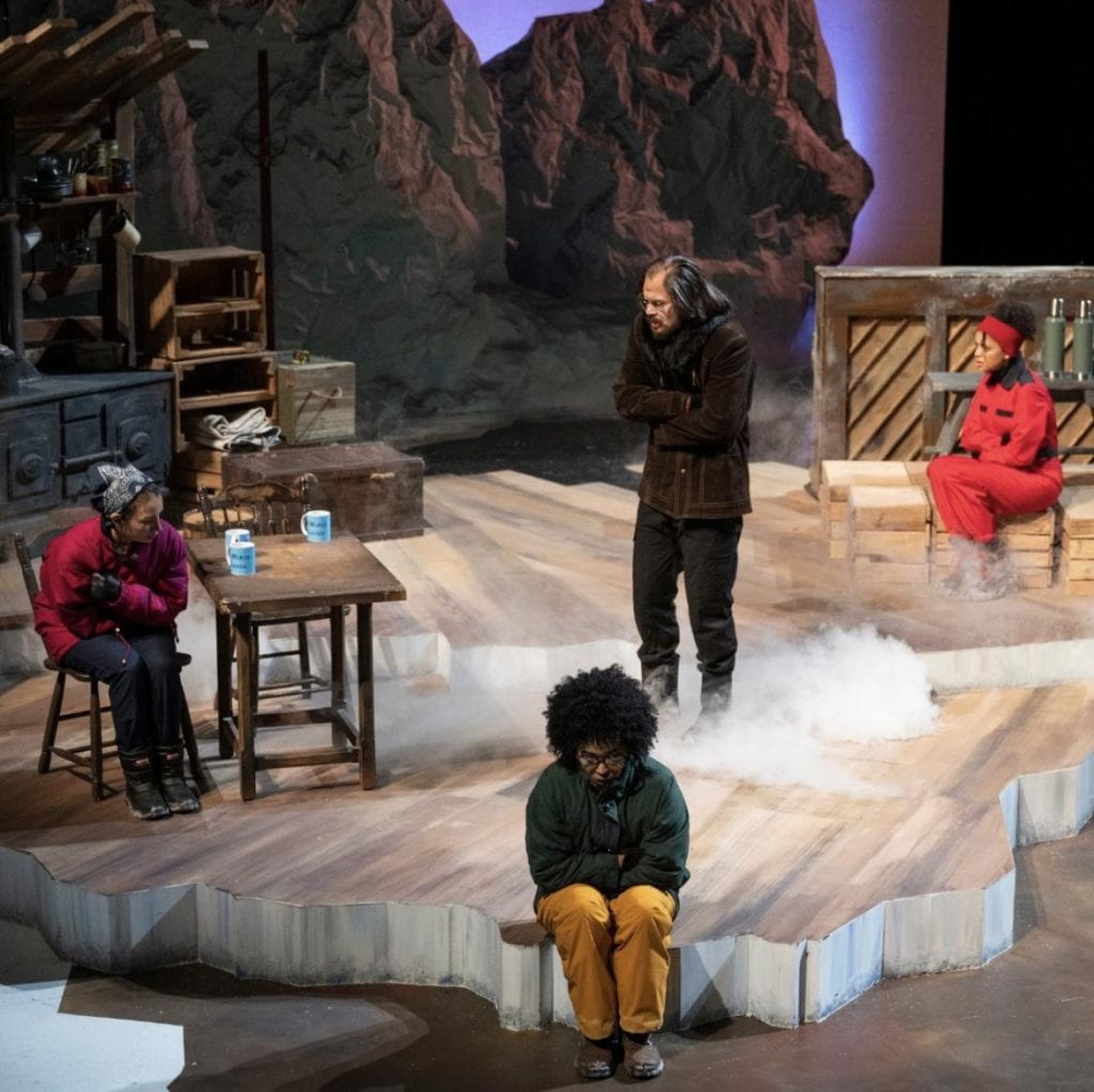 Four DePaul students on a stage in various positions on a wooden platform and chairs.
