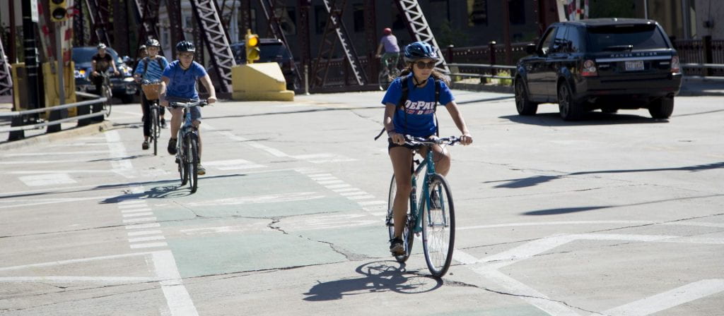 Three bicyclists wearing helmets and DePaul T-shirts ride through a sunny intersection.
