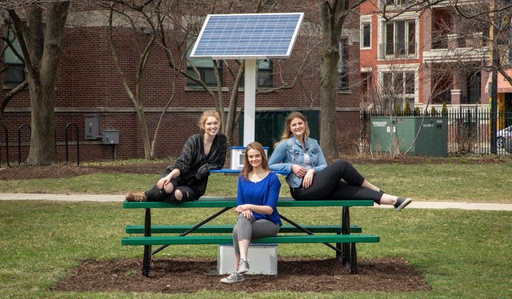 Three students sit at a green picnic table at DePaul's Lincoln Park campus. Above the table is a small solar panel attached to a metal pole. 