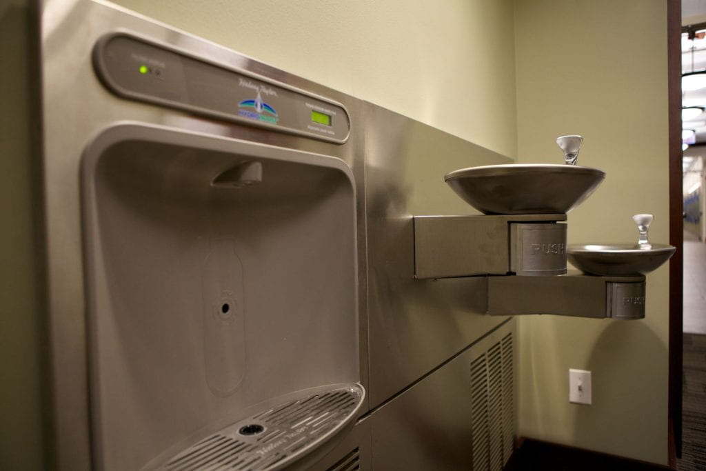 A silver water bottle filling station next to a water fountain in a DePaul campus hallway.