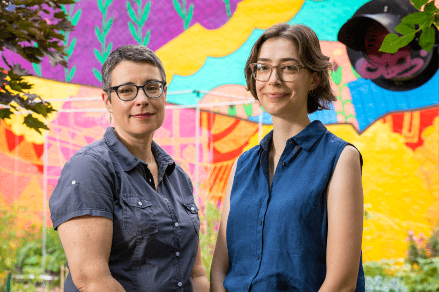 Faculty members Win Curran, a white woman wearing short hair, glasses, and a button-up shirt and Michelle Stuhlmacher, a white woman wearing medium-length hair, glasses, and a sleeveless blouse, in front of a colorful abstract mural. 
