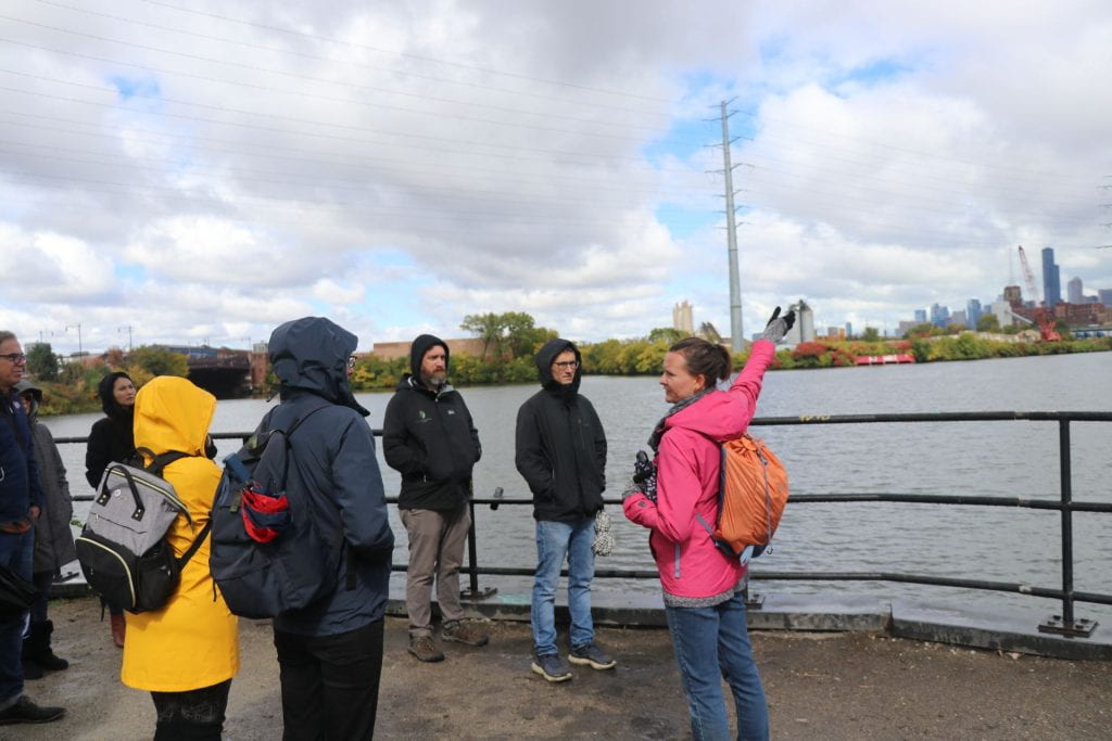 Becky Lyons, a white woman in a pink jacket and jeans, points to the river overlooking part of Chicago's skyline. A small group of students stands around her, facing the river.